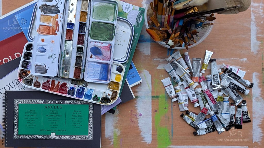 Learn to draw before you paint. Flat lay of expensive beginner art mistakes including Arches watercolor blocks, QoR and Da Vinci paint tubes, and a messy palette.